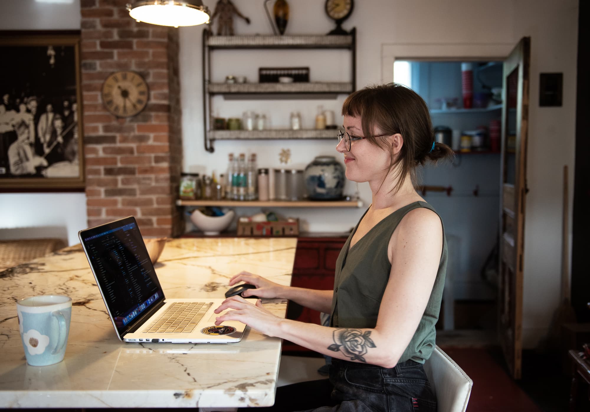 A woman with glasses and a tattoo on her arm is working on a laptop at a kitchen counter, smiling as she browses About Riley I Design. A mug sits nearby in the cozy kitchen with brick walls and shelves in the background.